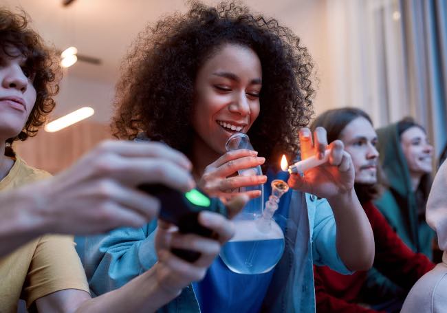 a young woman lights a bong filled with cannabis among her friends, sitting on a couch